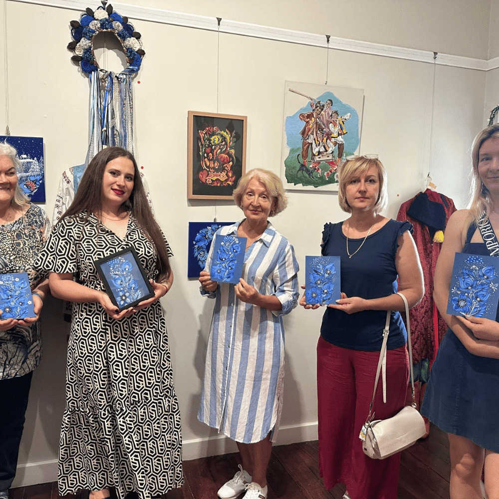 Five women stand side by side indoors, each holding a blue decorative book. Artworks and a floral wreath hang on the wall behind them. The women are dressed in summer clothes and facing the camera, smiling.