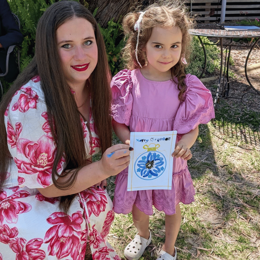 A woman and a young girl, both in pink floral dresses, pose outdoors. The woman holds a colorful, flower-themed drawing with "Happy Springtime" written on it. They are smiling and standing on grass near a tree.
