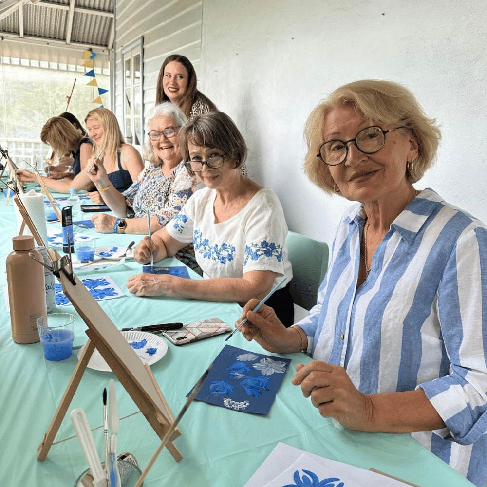 Six women sit at a long table covered with a light blue cloth, painting on canvases and smiling at the camera. Art supplies and blue-themed artwork are visible on the table in a bright, airy room.